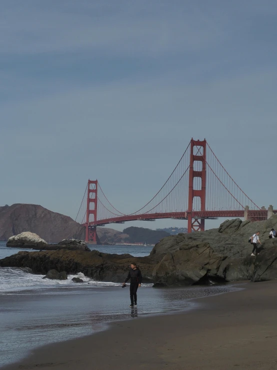 people are flying kites on the beach below a suspension bridge