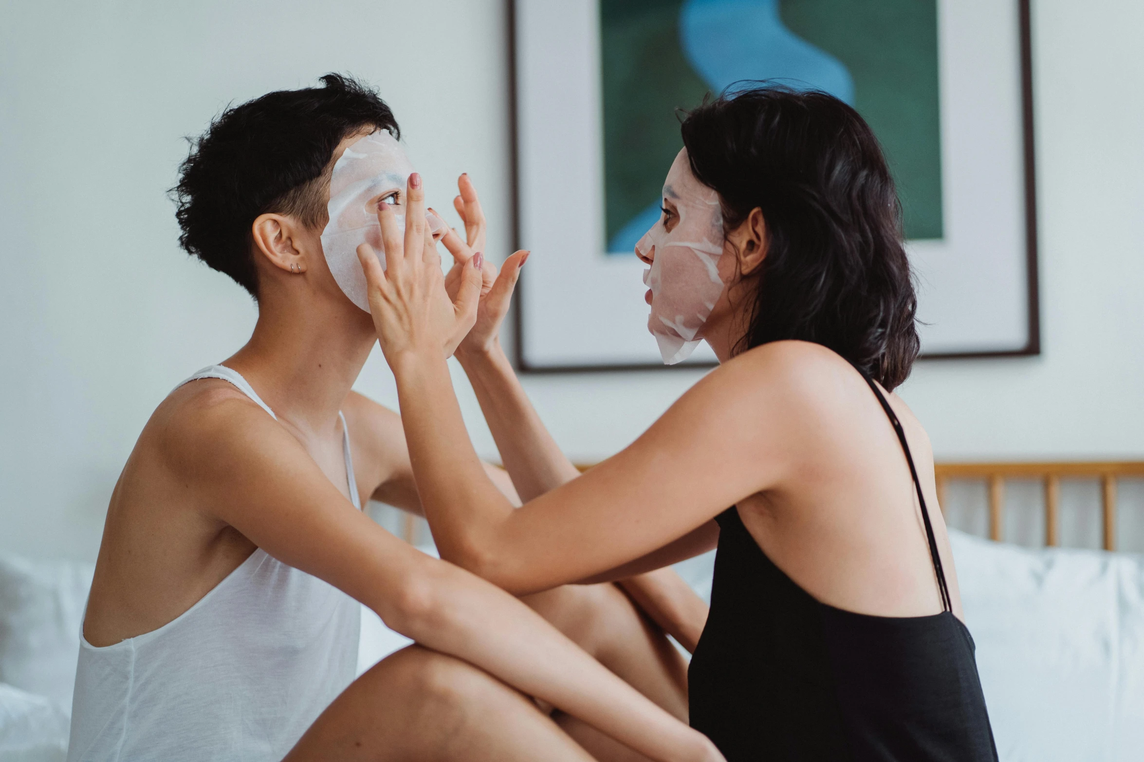 two women are sitting on the bed putting soing in their face