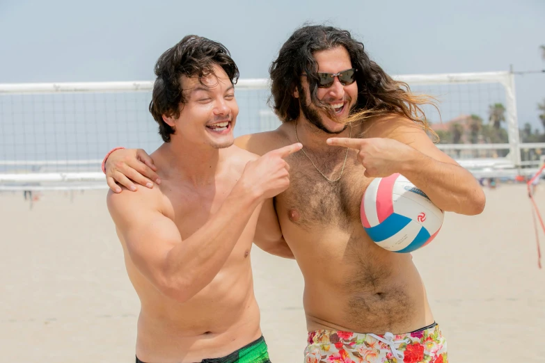 two men stand at the beach together as the man with a volleyball ball points to the viewer