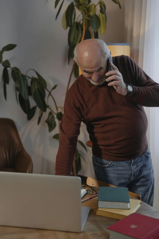 an old man standing at a table in front of his laptop while talking on a cell phone