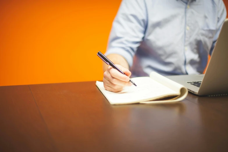 someone writing with a pen and notebook next to laptop