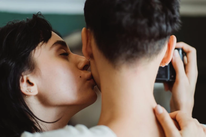 two people sharing a kiss while one woman is holding a cellphone