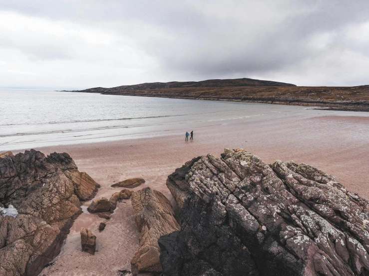 two people are walking along the shoreline near rocks