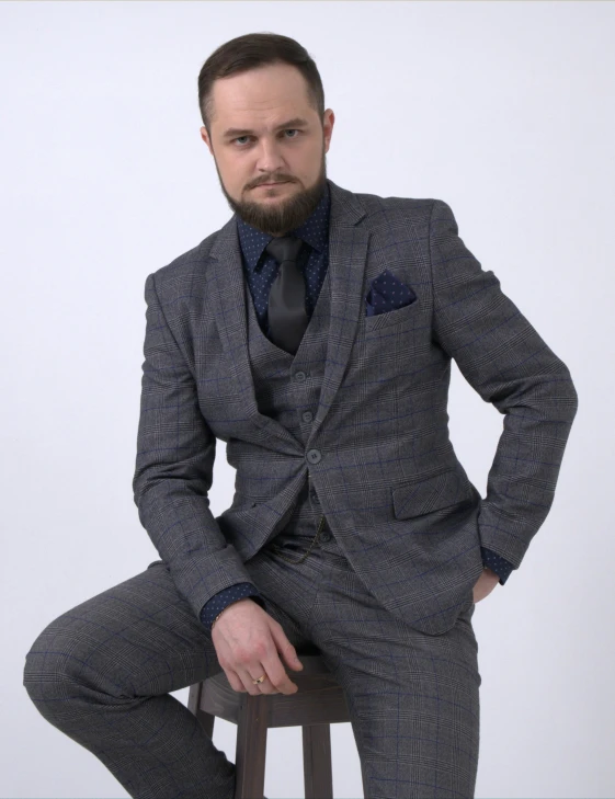 a young man wearing a grey and black suit, sitting on a stool