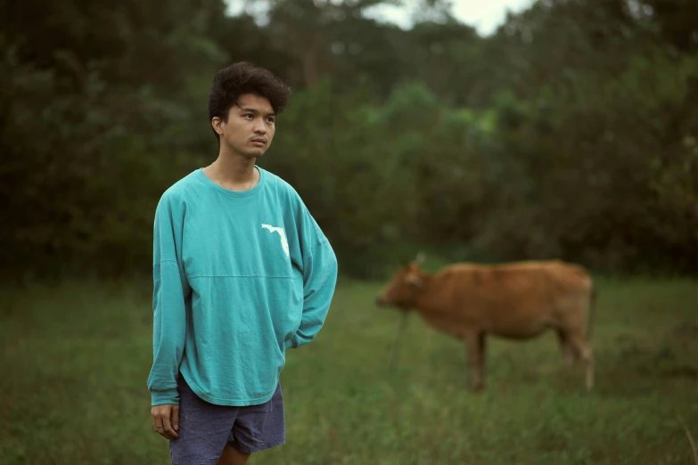 a boy stands with his hand on his hip as a cow walks by him