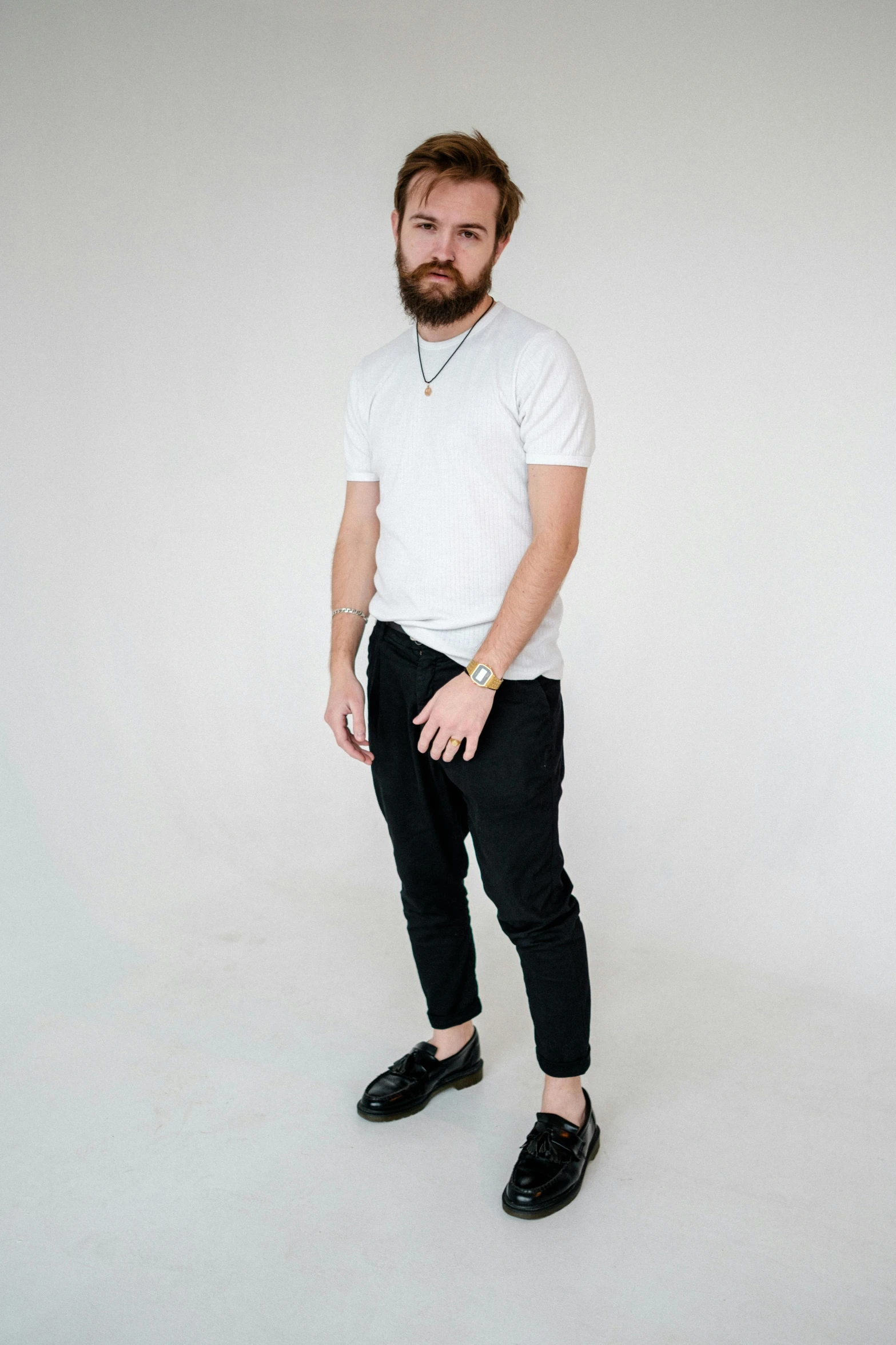 a bearded man in black pants and a white shirt is standing against a white wall
