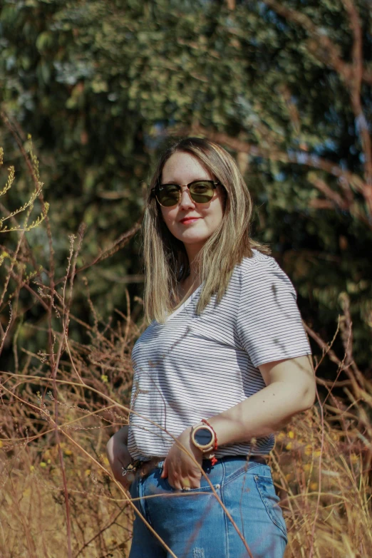 a woman stands in tall grass, wearing sunglasses