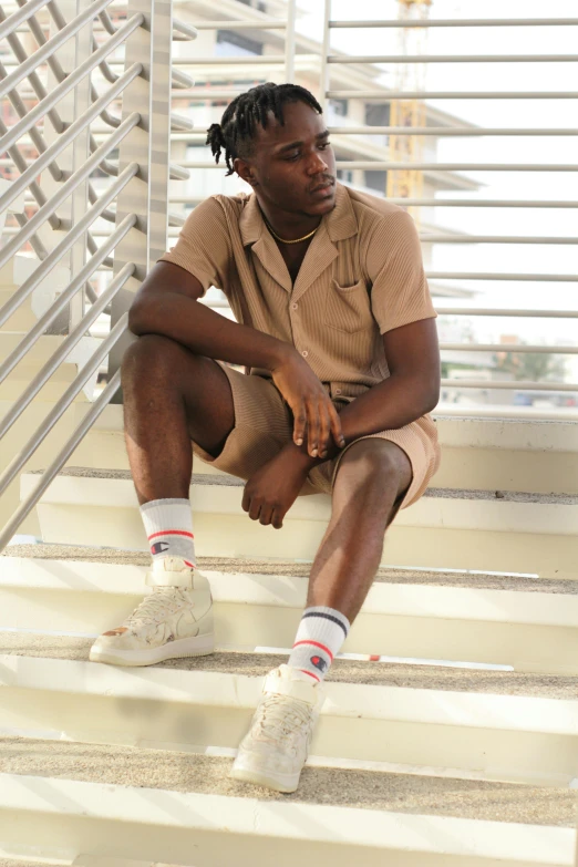 a young black man in a tan sweater sits on stairs looking out the window