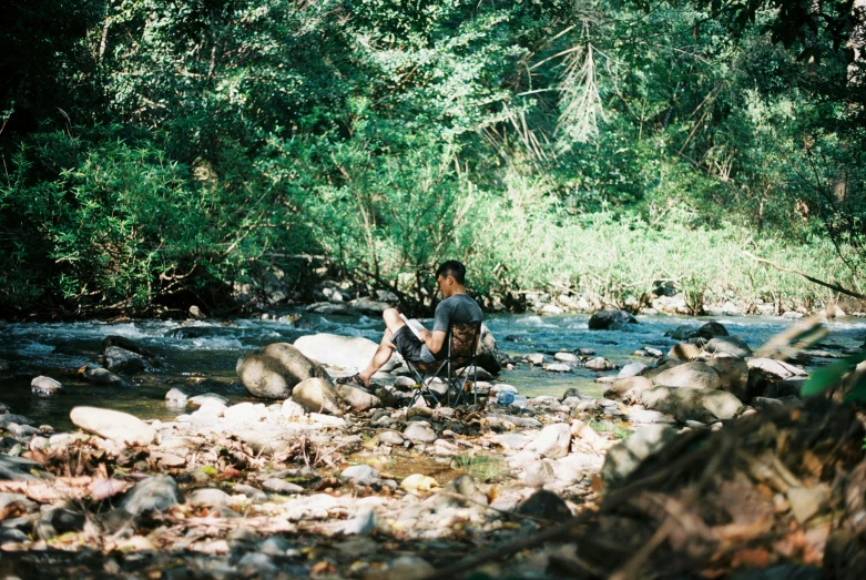 a man sitting on the bank of a stream near a forest