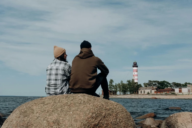 two people sit on a rock facing the ocean and a light house is in the background