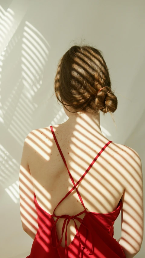 a woman wearing a red dress with a back cut, is standing near a wall with her hair pulled back