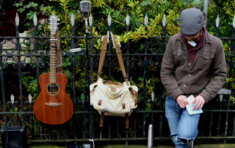 the man is sitting on the sidewalk near a fence, next to two guitar's that are hanging up against a black iron fence