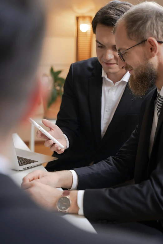 two men in suits sitting at a desk looking at a cell phone