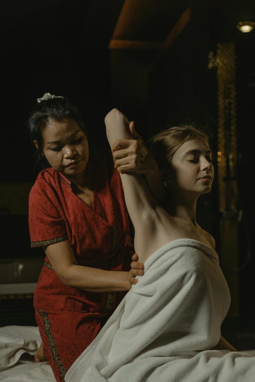 a woman sitting on a bed with another person getting her hair cut