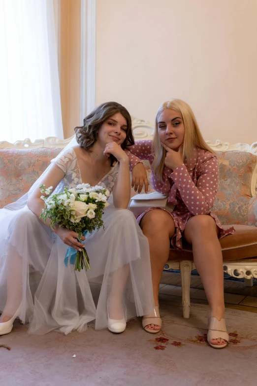 two woman pose together on a couch in white outfits