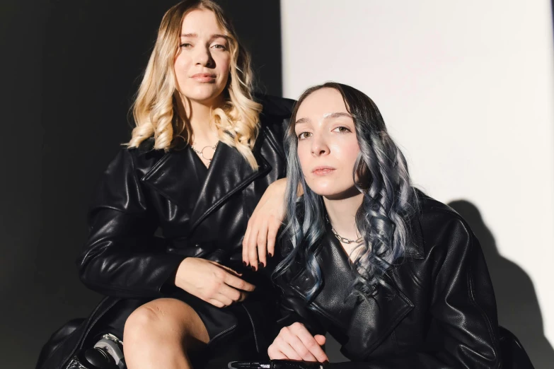 two women sitting next to each other, one of them is wearing a leather jacket and the other has her arms around her body