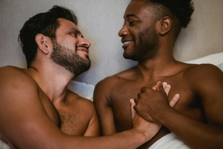 two young men in bed with white sheets one is rubbing his chest while the other is laughing