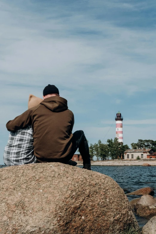 two people sitting on rocks at the edge of the water looking at a lighthouse