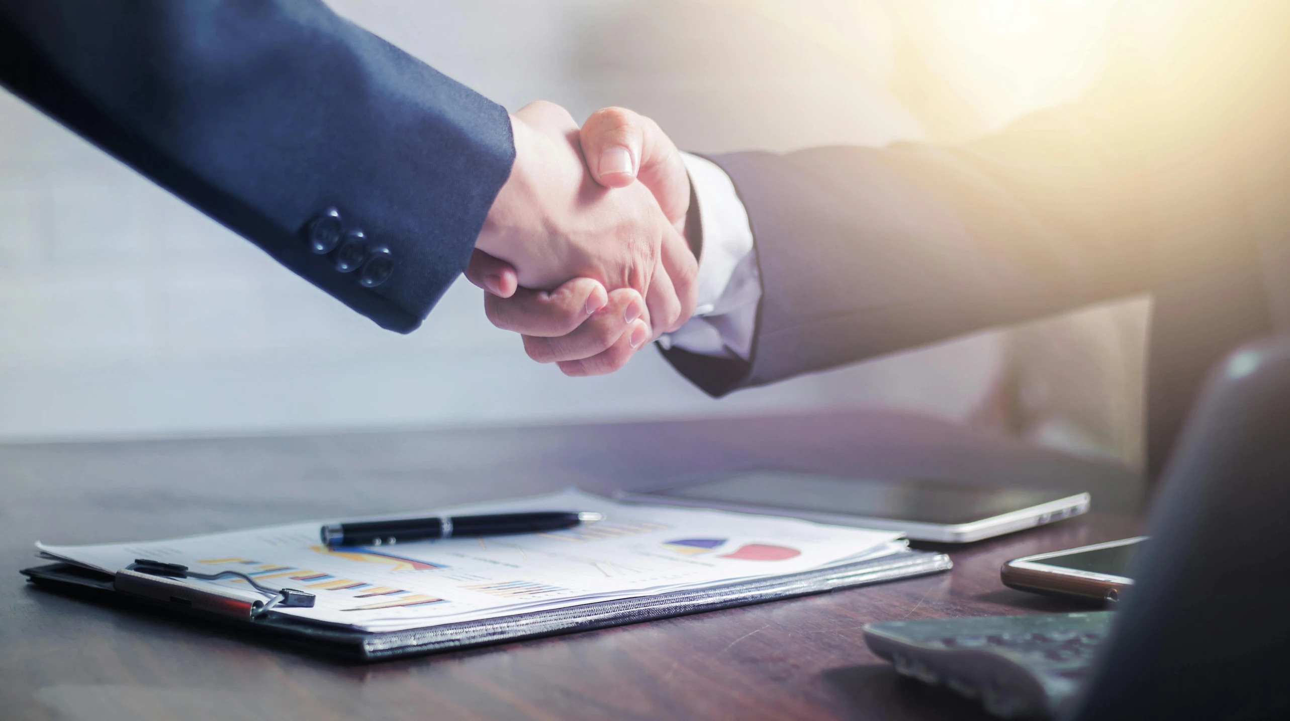 two people shaking hands over some papers and pens on a table