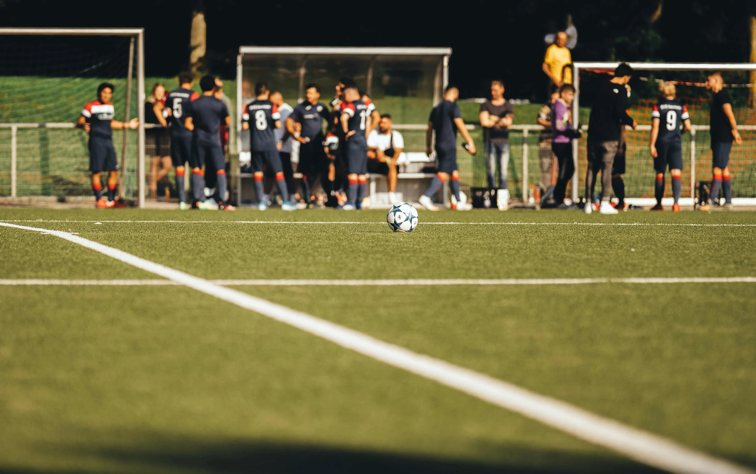 several people stand at the sideline as a soccer ball sits on a field