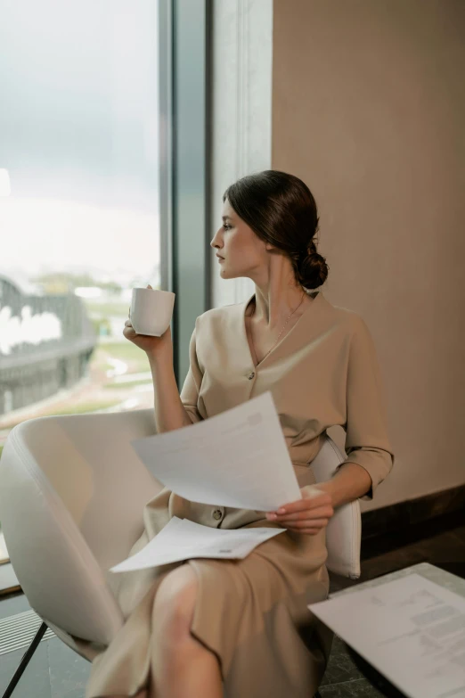 a woman sits near a window and looks out the window, holding a cup of coffee