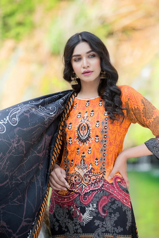 a young woman poses in front of the camera with her hand on her hip, wearing an orange and black shirt and large earrings