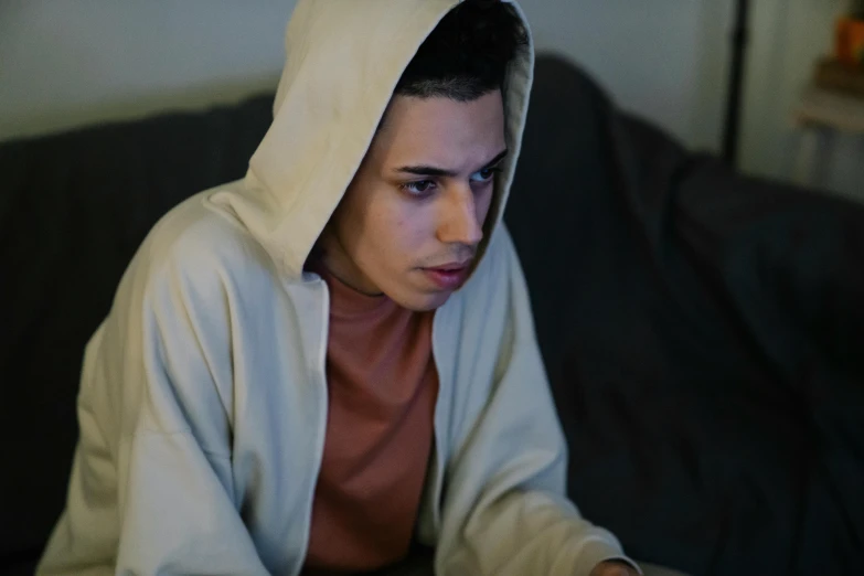 a man with a hoodie sitting on a couch using a laptop