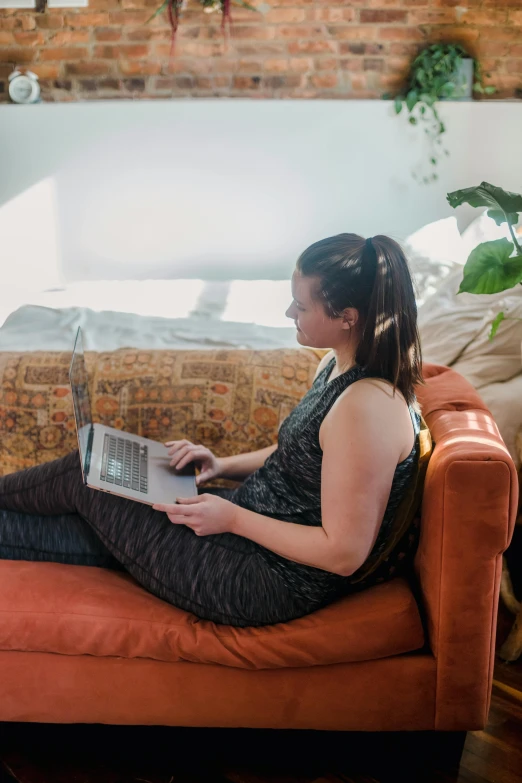 woman sitting on orange couch with laptop in front of brick wall