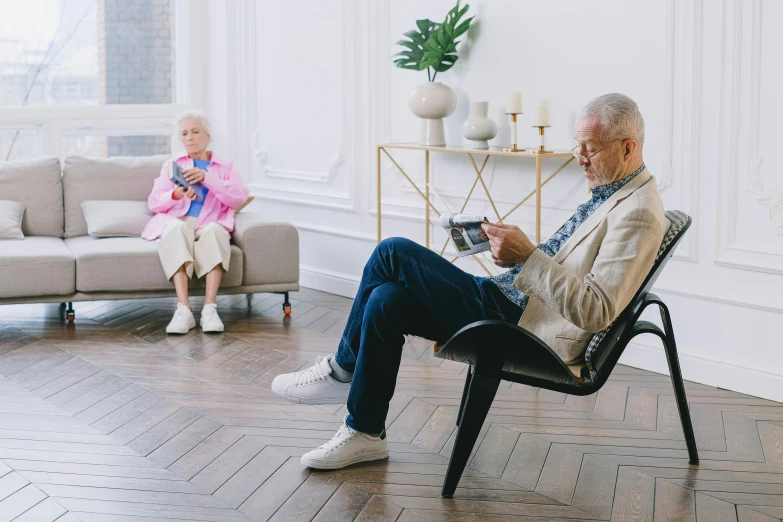 an older man sits in a chair holding a book and looks at another person