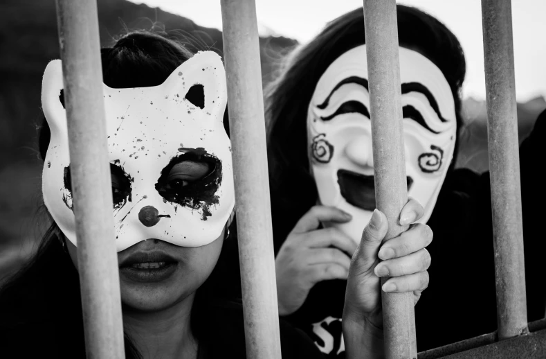 two women behind bars, one holding on to the mask of the other