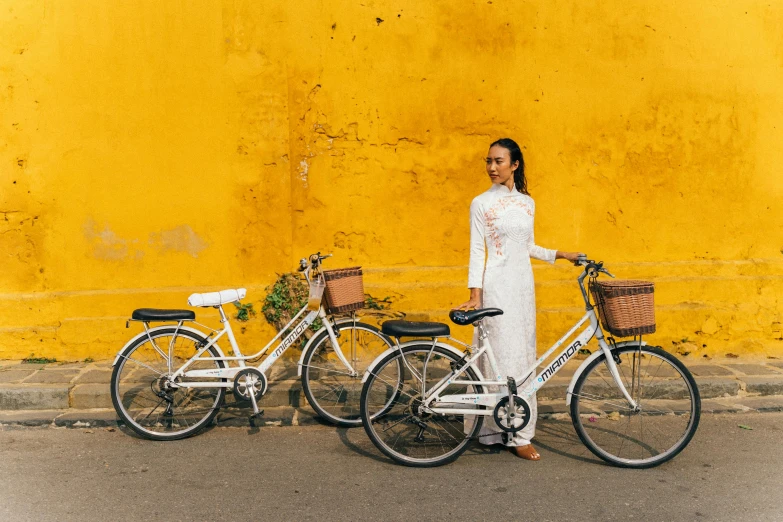 two white bicycles and a woman posing by a yellow wall