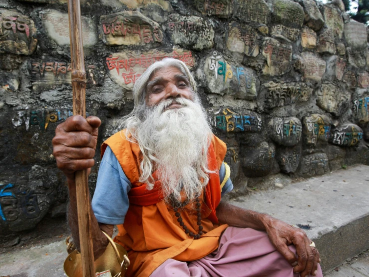 a man with white hair and beard sits in front of a stone wall