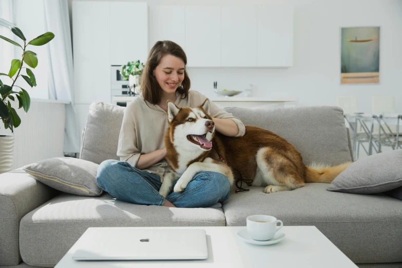 a woman sits on the couch with her dog while smiling