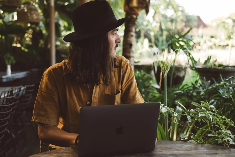 the man wearing a fedora looks off to his right as he uses a laptop in a cafe