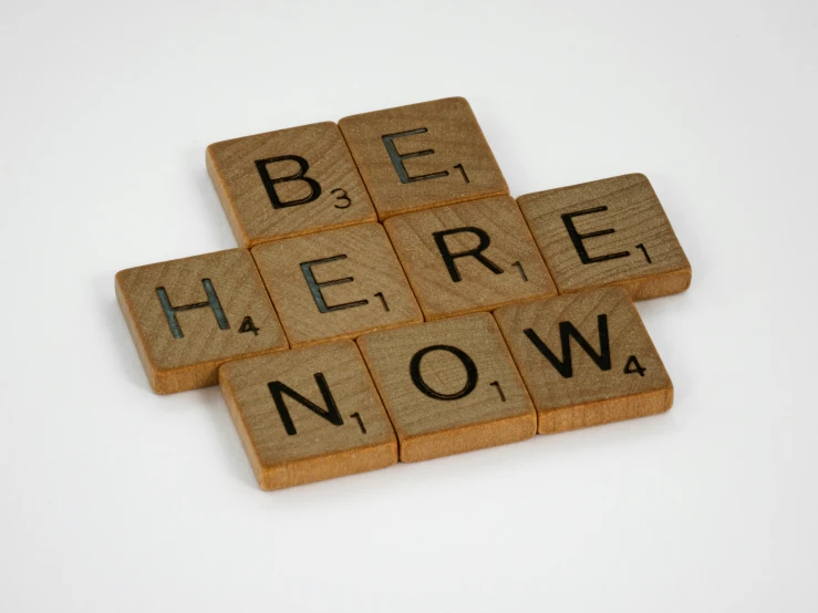 a close up of a wooden scrabble block with the words be here now on it