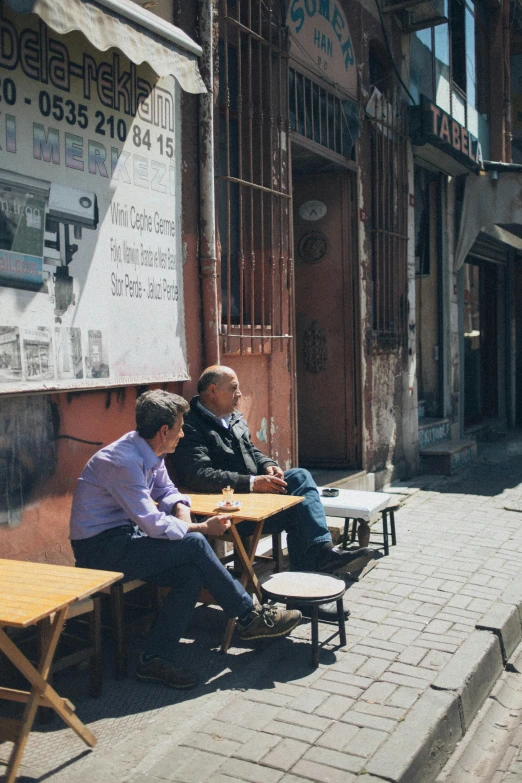 two men sitting on a bench with tables outside of a building