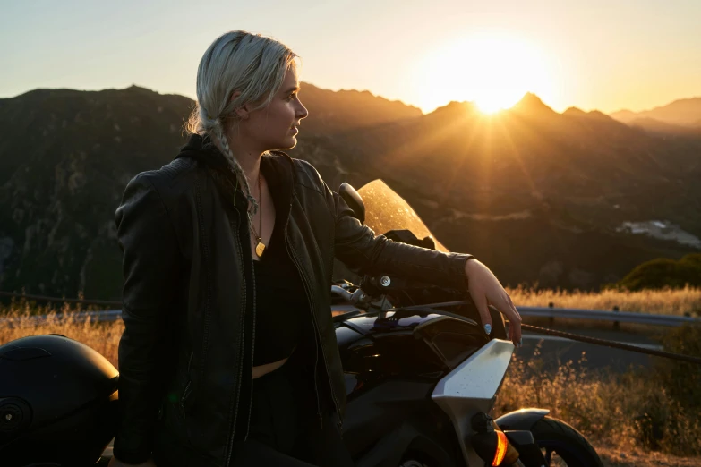 a woman standing next to her motorcycle in the sunset