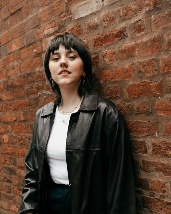 a woman in a leather jacket leaning on a brick wall