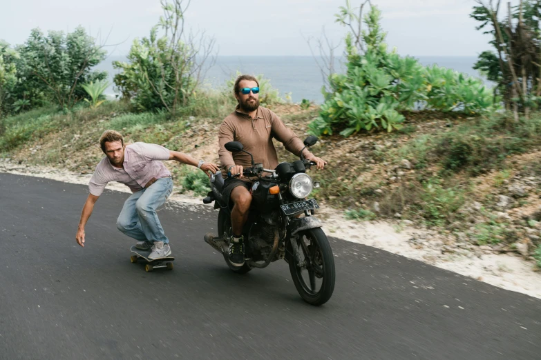 man on motorcycle with skateboard leaning over another guy