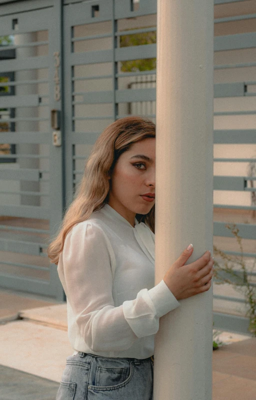 a woman wearing a white top and denim jeans leaning against a pole