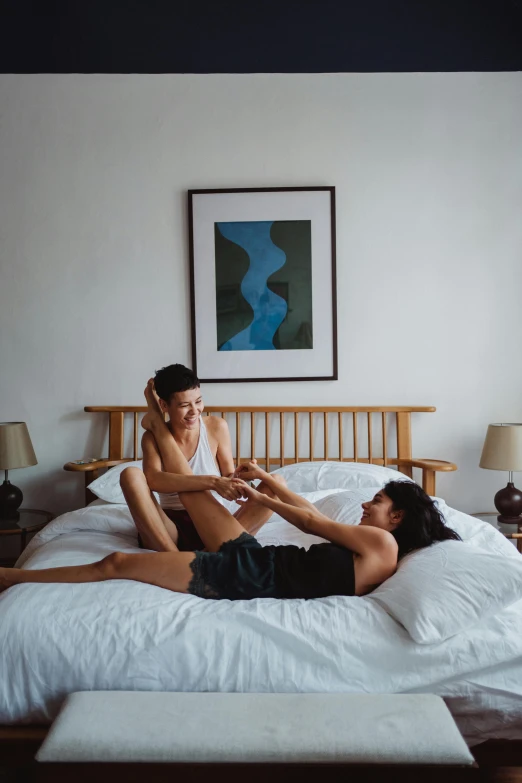 two women sit on a bed, one stretching her leg, the other falling asleep