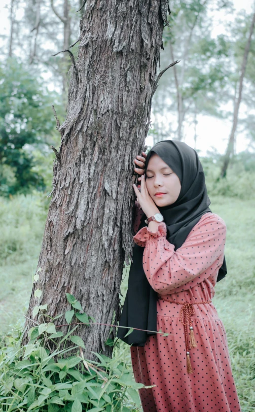 a woman is posing by a tree on a cell phone