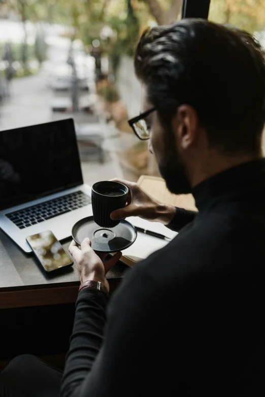 a man with a coffee cup in his hand sitting at a table next to a laptop