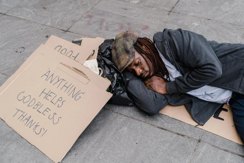 a homeless man sleeps on a cardboard box with handwritten sign