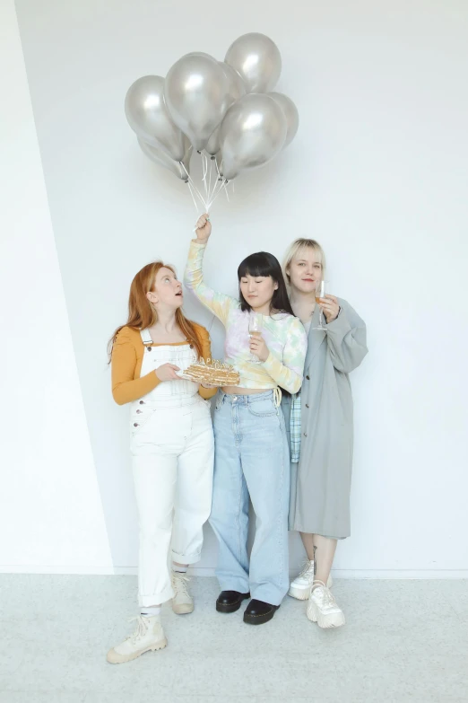 three young women holding balloons, standing in front of white wall