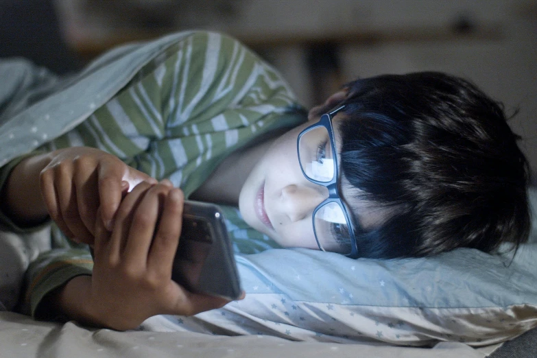 the boy wearing glasses is laying in bed and reading a book