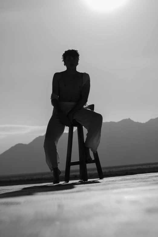 a black and white image of a person sitting on a stool with mountains in the background