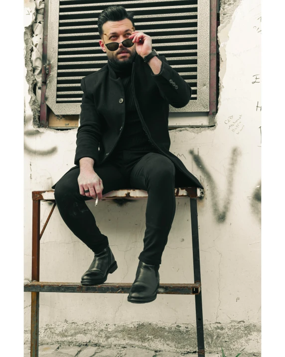 man with a black suit and tie, sitting on a chair, smoking a cigarette