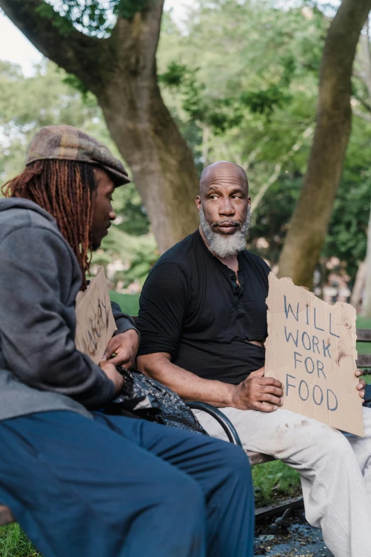 two men sit on a bench, one with a sign saying will work for food