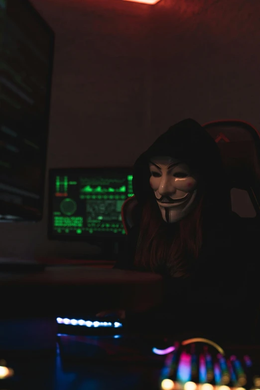 a person is sitting in a dark room, wearing a black mask and using their computer